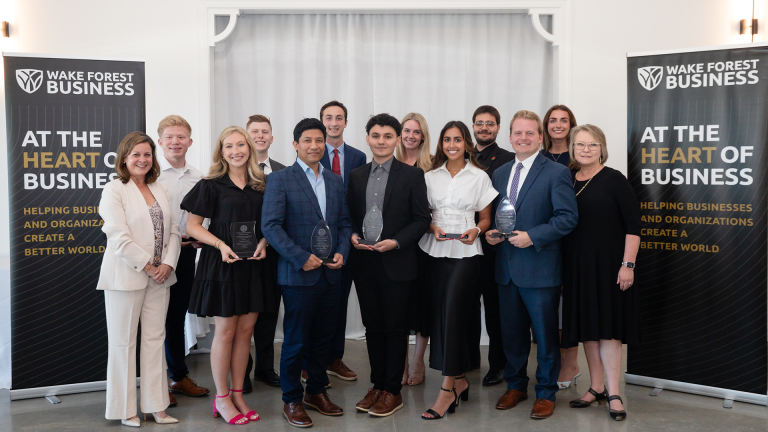 A group of diverse students and faculty, some holding awards, stand in front of two "Wake Forest Business: At the Heart of Business" banners.