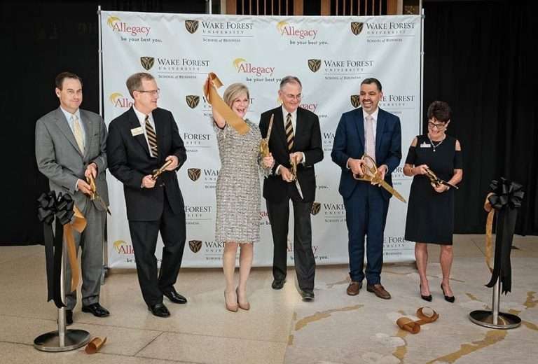 Six individuals, including representatives from Allegacy and the school, holding large golden scissors during a ribbon-cutting ceremony to celebrate renaming.