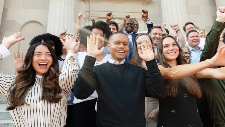 A diverse group of smiling business students and professionals standing on the steps of a university building, cheering with raised hands and fists.