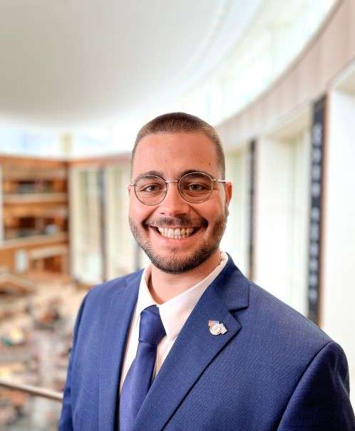 A close-up headshot of a person with short dark hair, glasses, and a beard, smiling and wearing a blue suit jacket over a white shirt.