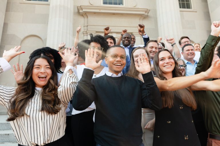 A diverse group of approximately 15 to 20 people stand on stone steps in front of a classical building with large columns. They are smiling and cheering enthusiastically; several people in the front have their hands raised in waves, while those in the back have their fists raised in celebration. The group is dressed in business-casual attire, including sweaters, button-down shirts, and dresses. The lighting is bright and sunny.