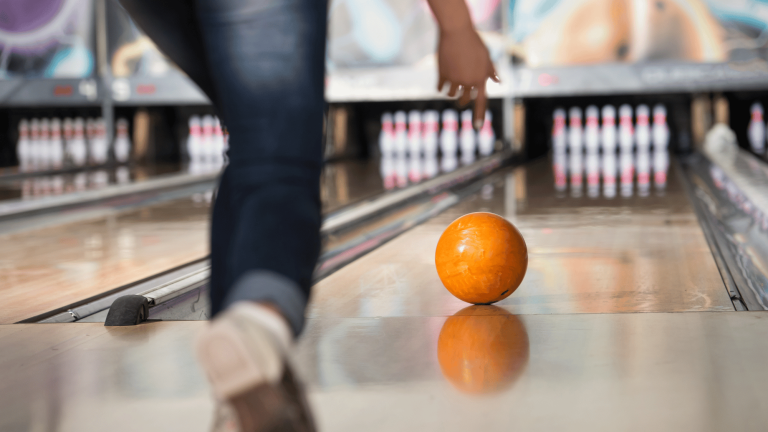 An orange bowling ball rolls down a polished wooden lane toward a set of pins.