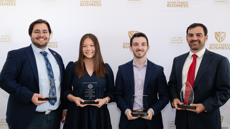 Four individuals stand in front of a white backdrop with the Wake Forest Business logo, each holding an award and smiling.