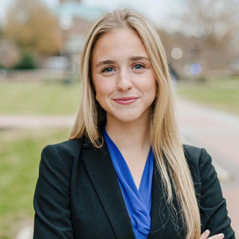 A person wearing a black blazer over a blue shirt stands with their arms crossed, looking forward with a slight smile. They have long blonde hair and are outdoors in front of a blurred background of trees and buildings.