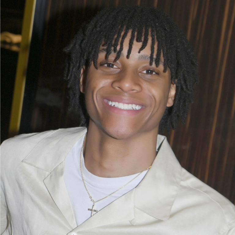 A young person with dreadlocks smiles at the camera, wearing a light-colored beige shirt over a white shirt.