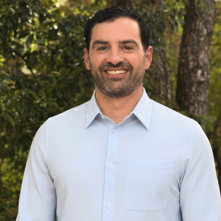 A man with dark hair and a beard, wearing a light blue collared shirt, smiles at the camera. He is outdoors with trees and foliage in the background.