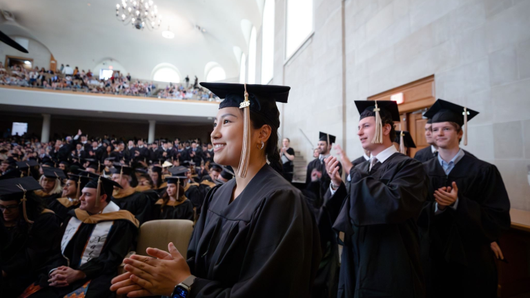 A smiling graduate in a black cap and gown claps during a business school hooding ceremony in a crowded auditorium, with other graduates applauding in the background.