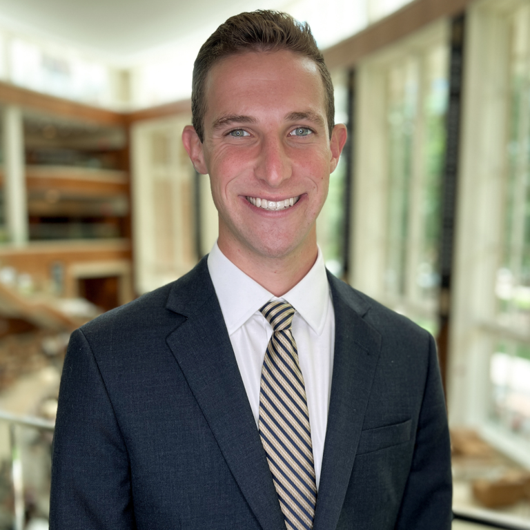 A man in a dark suit and striped tie smiles at the camera in front of a blurred background of a room.