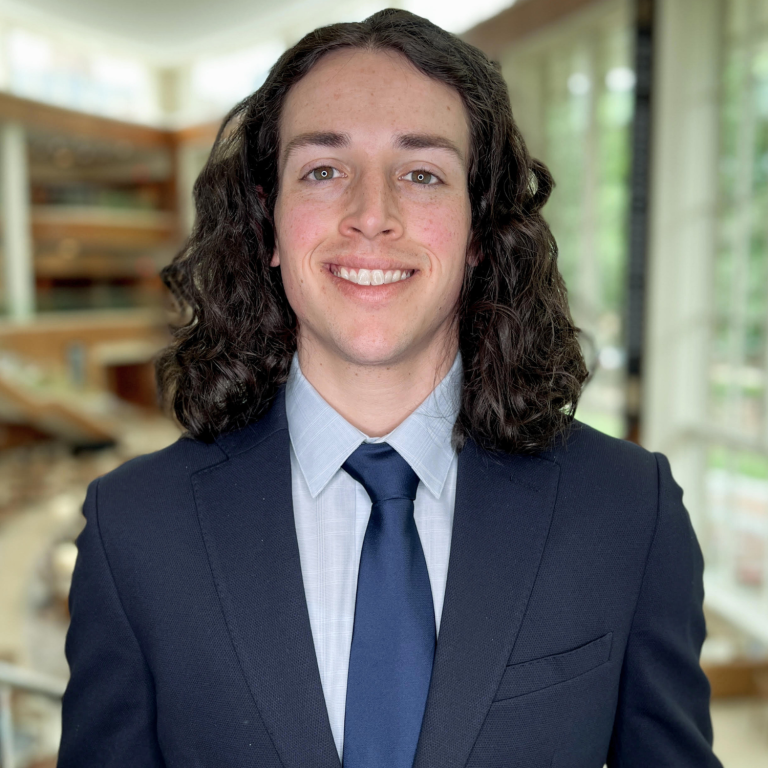 A person with curly dark hair wearing a navy suit and a blue tie smiles at the camera in front of a blurred background of a building interior.