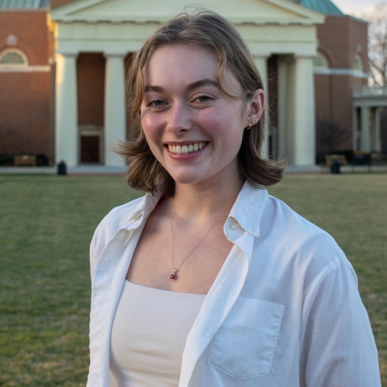 A person with curly hair and glasses smiles at the camera, wearing a white unbuttoned shirt over a beige top. Behind them is a building with large columns and a grassy area.