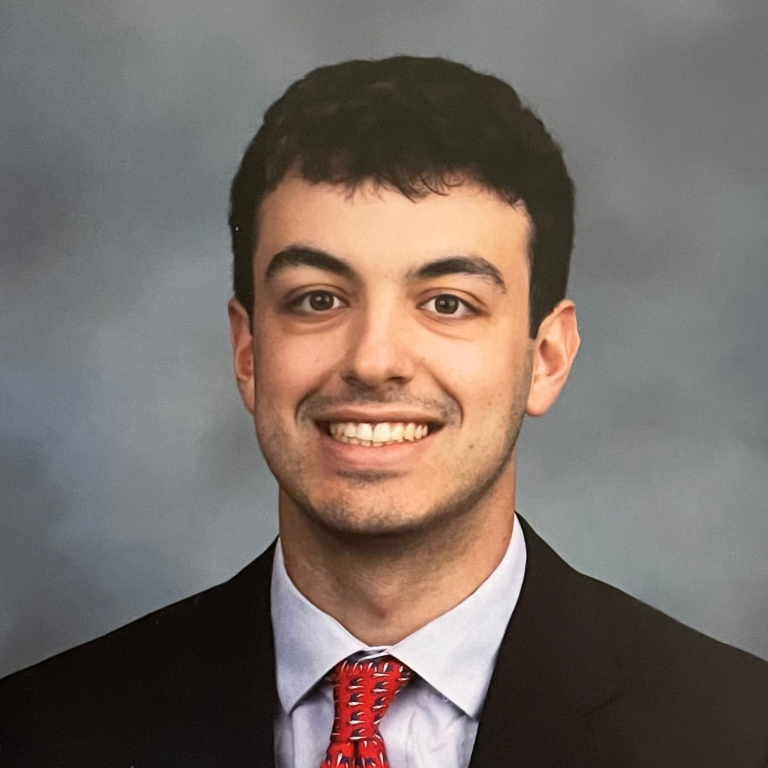 A headshot of a person wearing a suit jacket, white shirt, and red tie against a gray background.