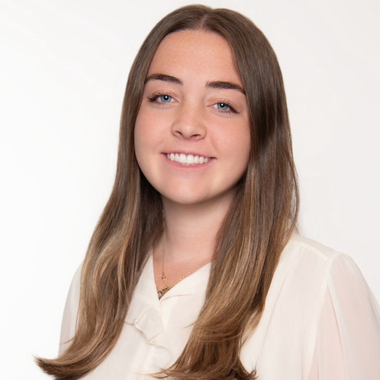 A young person with long brown hair and blue eyes smiles at the camera, wearing a light-colored blouse against a white background.