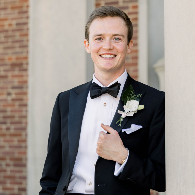 A man in a tuxedo with a bow tie and a boutonnière stands with his hand on his lapel, with a brick wall and a column in the background.