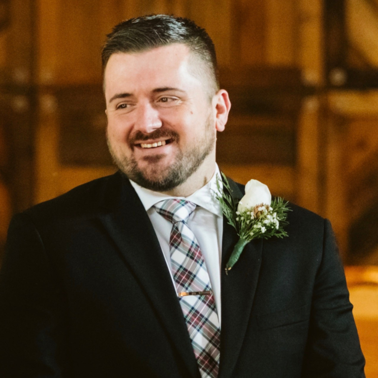 A man in a suit and tie smiles with a boutonniere on his lapel.