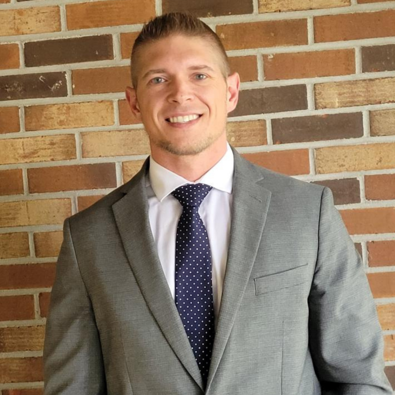 A man wearing a grey suit, white shirt, and blue tie stands in front of a brick wall.