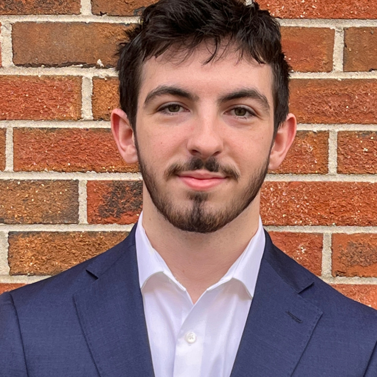 A close-up headshot of an individual wearing a dark blue suit and a white collared shirt against a brick wall background.