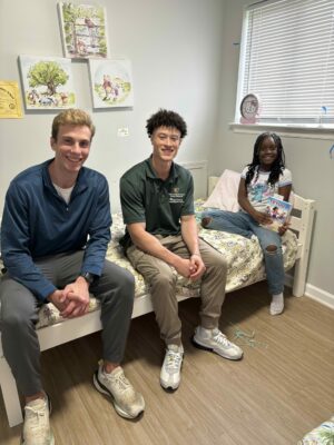 Two ACLC Scholars sit with a child beside a newly delivered bed as the child holds a book.